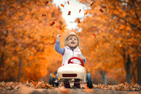 Young child playing on toy car surrounded by orange leaves and trees, smiling